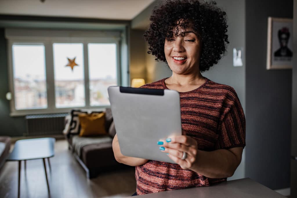 Woman with curly hair smiling at a tablet in a cozy living room. Natural light from large windows, star decoration visible outside, serene atmosphere.