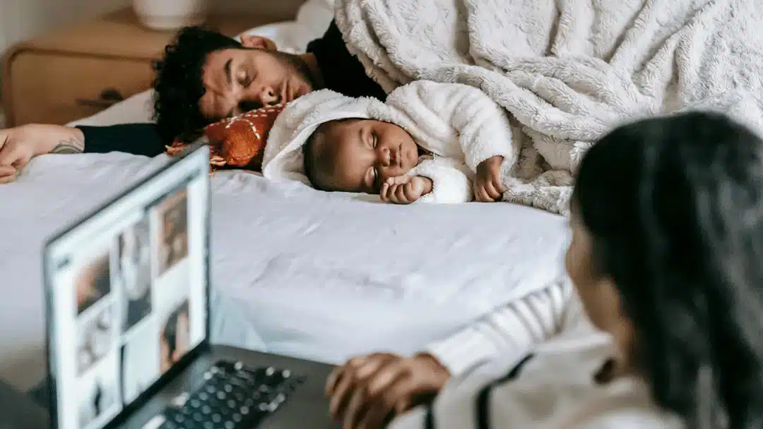 A person using a laptop sits on the edge of a bed, while another person and a baby dressed in a white outfit relax under a fluffy blanket in the background.