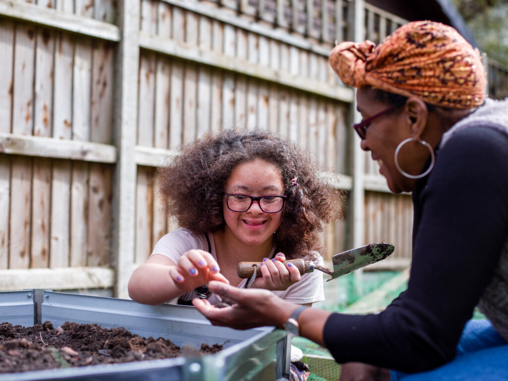 A young girl with glasses digs in a garden bed, smiling at a woman handing her a seed. They are enjoying the day in a fenced backyard.