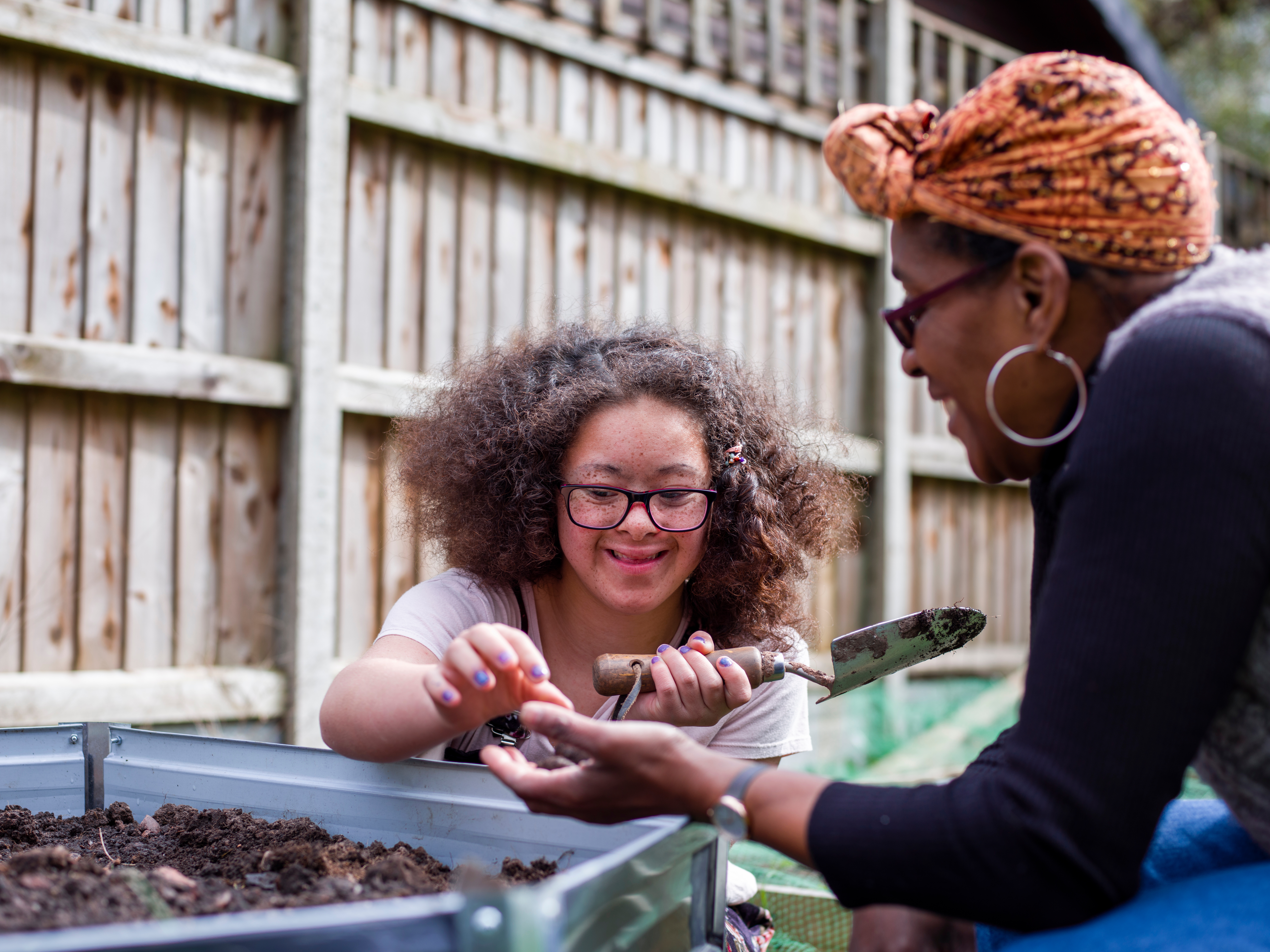 A young girl with glasses digs in a garden bed, smiling at a woman handing her a seed. They are enjoying the day in a fenced backyard.