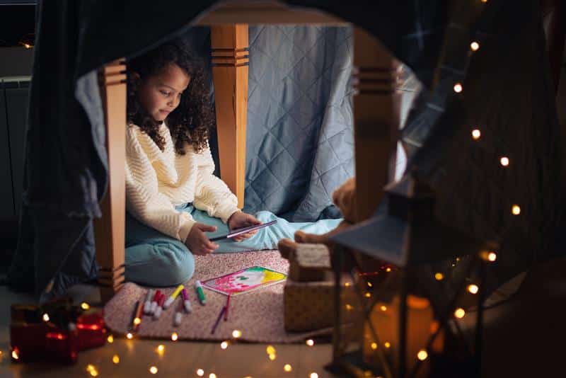 Foster care child sitting under a table in a cozy fort, drawing on paper with markers, surrounded by warm lights and toys.