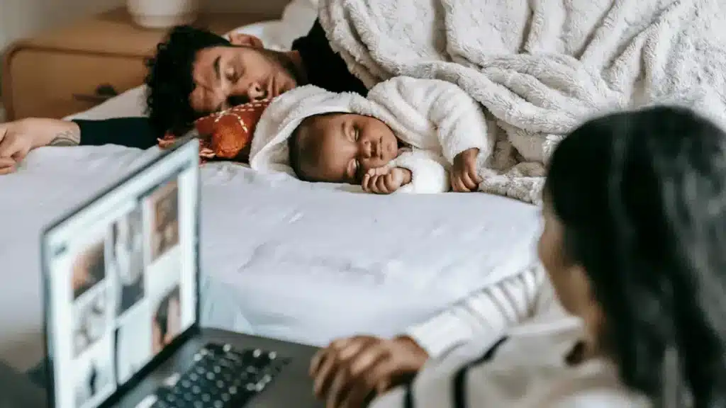 A person using a laptop sits on the edge of a bed, while another person and a baby dressed in a white outfit relax under a fluffy blanket in the background.