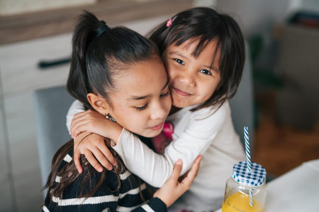 Two young girls share a warm hug. A jar of juice is nearby at a table indoors. 